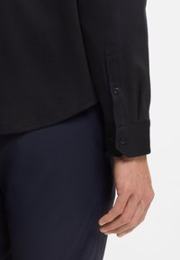 Μαύρο πουκάμισο με μανσέτα με κουμπιά, σχεδιασμένη με αιχμηρή μανσέτα και λεία, βαμβακερή υφή. Φορεμένο πάνω από σκούρο μπλε παντελόνι.