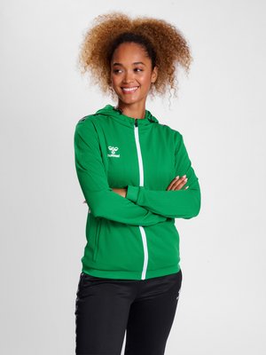 Mujer con cabello rizado sonríe mirando de lado, vestida con una sudadera con capucha verde y pantalones negros con logo. Brazos cruzados.