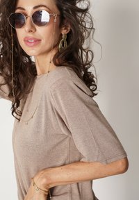 Kobieta z kręconymi włosami, ubrana w beżową bluzkę, złote kolczyki, bransoletkę z zawieszką w kształcie serca i okulary przeciwsłoneczne z złotym łańcuszkiem na tle jednolitym.