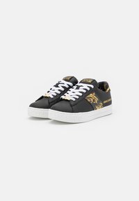Black low-top sneakers with white laces and soles, gold baroque pattern on sides, and "Versace Jeans Couture" branding on tongue and heel.