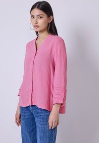 Chemise rose à col V et manches trois quarts avec des accents plissés texturés sur les manches. Portée avec un jean bleu.