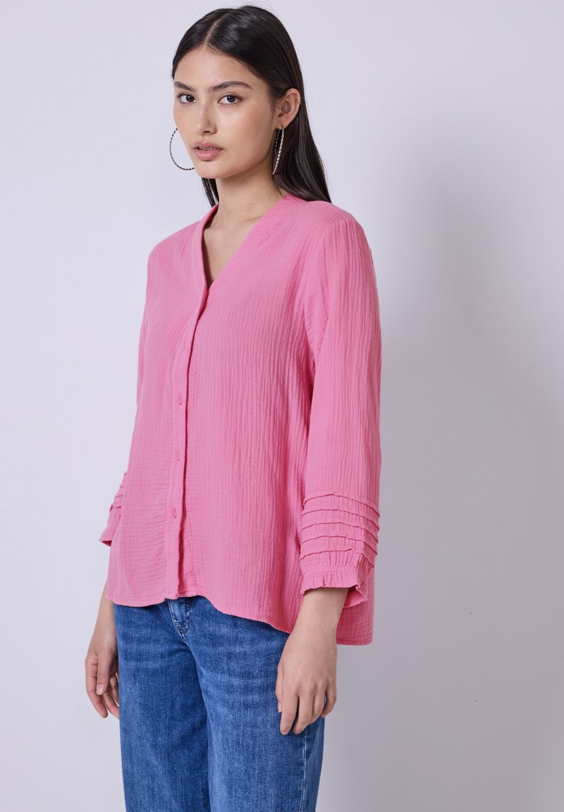 Chemise rose à col V et manches trois quarts avec des accents plissés texturés sur les manches. Portée avec un jean bleu.