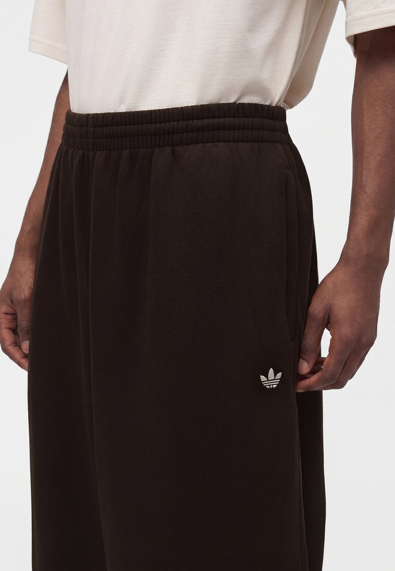 Mand iført sorte Adidas joggingbukser med elastisk talje og lille hvidt Adidas-logo på venstre lår, kombineret med en hvid skjorte.