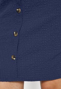 Detailní záběr na tmavě modrou texturovanou sukni s třemi tlačítky z želvoviny na přední straně a viditelnou boční kapsou.