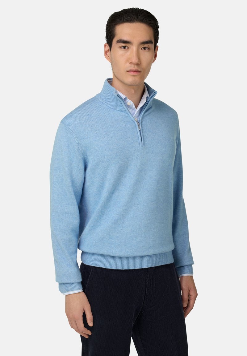 Pull à col zippé bleu clair en tissu doux, avec des poignets et un ourlet côtelés. Porté par-dessus une chemise blanche à rayures, présenté sur un mannequin.