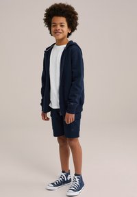 Veste à zip marine avec capuche ajustable, t-shirt blanc, shorts cargo marins et baskets montantes avec semelles blanches. Tenue décontractée.