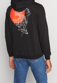 Sudadera negra con un dibujo en línea blanca en la parte posterior, que representa una cara y cabello contra un gran círculo rojo. Tejido suave, corte relajado.