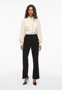 Kvinne med krøller som har på seg en kremfarget bluse med rysjer, svarte tettsittende bukser og svarte spisse høye hæler, som står foran en hvit bakgrunn.