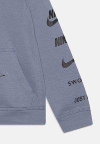 Šedá mikina Nike s černými logy. Obsahuje klokaní kapsu, ribbed manžety a hladkou, měkkou texturu. Design zahrnuje potisk textu na rukávech.