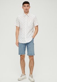 Camisa blanca de botón de manga corta con pequeño estampado floral azul y rojo, combinada con pantalones cortos de mezclilla azul claro y zapatillas blancas.