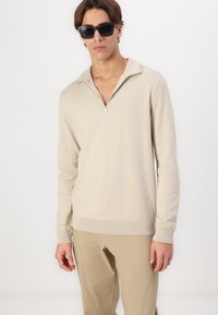 Suéter beige de cremallera en un material de punto suave, con mangas largas y puños acanalados, combinado con pantalones de color claro. Unas gafas de sol negras complementan el look.