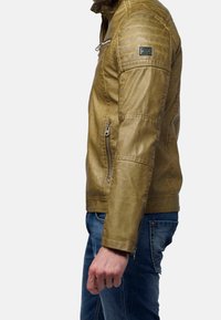 Giubbotto in pelle marrone con tasche con zip, dettagli in costina sulle spalle e finitura testurizzata. Indossato su jeans in denim blu.