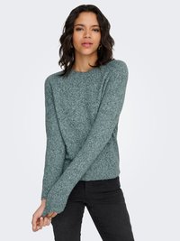 Naine kannab pehmet rohelist kirju kootud kampsunit ja musti pükse, tal on lõdvestunud kehahoiak ning käed on õrnalt rinnal risti.