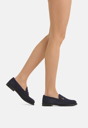 Gambe che indossano calze velate color nudo e mocassini in camoscio blu navy su uno sfondo chiaro uniforme.
