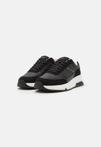 Baskets noires avec un mélange de matériaux lisses et texturés, dotées d'une semelle intermédiaire blanche et de perforations discrètes. Logo de la marque sur le côté.