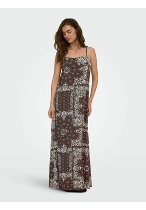 Kobieta w brązowej sukience maxi z białymi wzorami paisley i kwiatowymi patchworkami, stoi z zamkniętymi oczami i zrelaksowaną postawą.