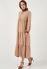 Beige dlouhé šaty s dlouhým rukávem, s vrstveným designem a texturovanou látkou. Obsahují tkanici s třásněmi a jsou doplněny hnědými sandály na podpatku.