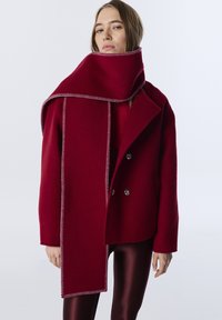 Červená bunda z vlněné směsi s širokým šátkem s kontrastním prošíváním, kombinovaná s lesklými burgundskými legínami. Dvouvrstvý přední díl.