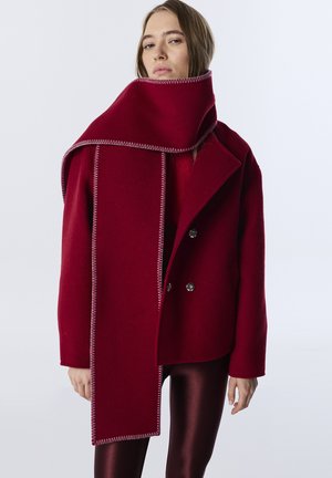 Rode wolmix jas met een brede sjaal met contrasterende stiksels, gecombineerd met glanzende bordeauxrode leggings. Dubbelgeknoopte voorkant.