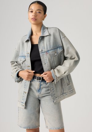 Kobieta nosząca oversize jasną kurtkę jeansową oraz dopasowane szorty jeansowe do kolan, czarny krótki top i czarny pasek.