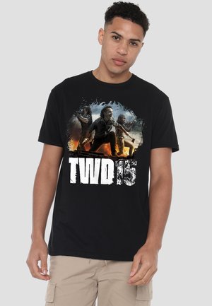 Junger Mann trägt ein schwarzes T-Shirt mit drei bewaffneten Figuren und dem Text "TWD 15" in Weiß auf der Vorderseite vor einem neutralen Hintergrund.