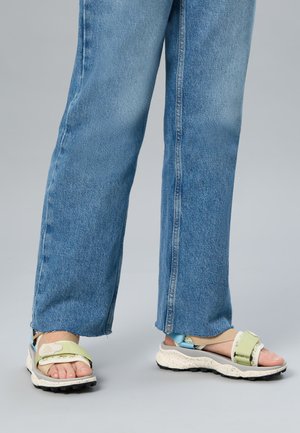 Personne portant un jean en denim bleu clair et des sandales épaisses ouvertes aux orteils avec des brides vert pastel et beige sur un fond gris.
