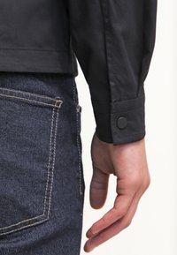 Lähedalt vaadates käsi tumeda denimpüksi tasku kõrval, kätetööl musta pika varrukaga särgi, millel on nööbitav mansett.