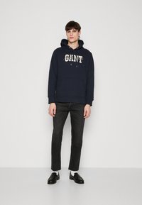 Felpa blu marina con logo "GANT", cappuccio con stringhe e tasca anteriore, abbinata a jeans neri slim-fit e scarpe nere.