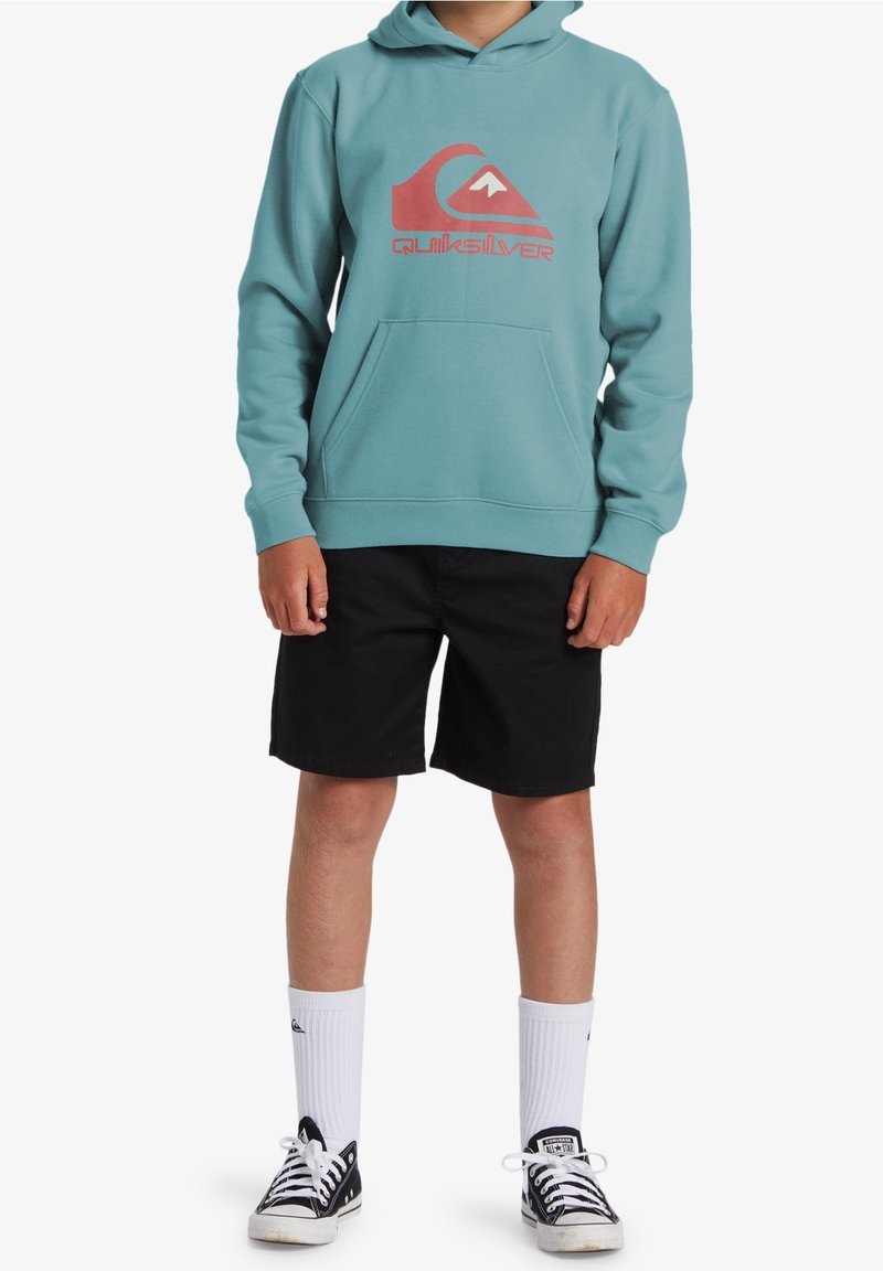 Personne portant un sweat à capuche bleu sarcelle avec un logo rouge, un short noir, des chaussettes blanches montantes et des baskets basses noires, debout devant un fond blanc.