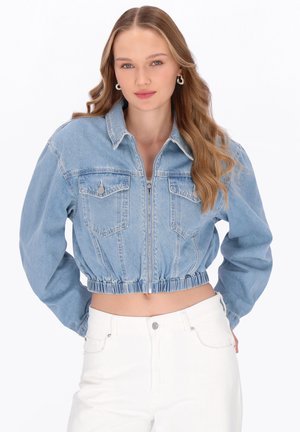 Fiatal nő hosszú hullámos hajjal, világoskék, rövidített farmerdzsekit felhúzva, fehér, magasított derekú nadrágban áll fehér háttér előtt.