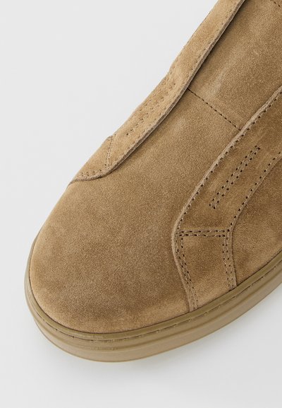 Close-up of tan suede shoe toe with detailed stitching and a rubber sole on a white background.