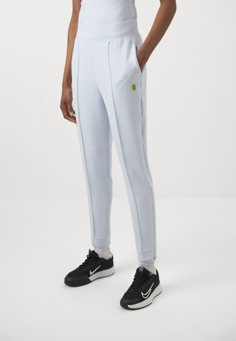 Hellblaue Jogginghose aus weichem Stoff, mit Vorderfalten, hoher Taille und einem kleinen grünen Logo. Kombiniert mit schwarzen Turnschuhen.