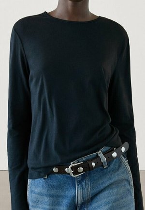 Persoon draagt een zwart shirt met lange mouwen, ingestopt in een blauwe spijkerbroek met een zwarte riem met zilveren studs en gesp.