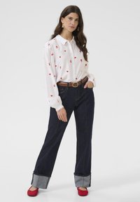 Bela majica z rdečimi srčkastimi vzorci, temno modre kavbojke z zavihanimi manšetami, rjav pas in rdeče balerinke. Dolgi rokavi z nabranimi manšetami.