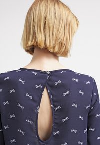 Blouse bleu marine avec imprimé de nœuds blancs, détail en ouverture dos, tissu doux, encolure arrondie et fermeture à boutons au dos.