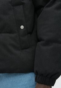 Schwarze gerippte Jacke mit Ärmel und Tasche, ausgestattet mit silbernem Druckknopf, getragen über hellblauen Denim-Jeans.
