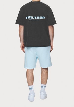 Uomo che indossa una maglietta nera oversize con il testo "PEGADOR", pantaloni corti azzurri, sneakers bianche Nike e calze bianche con scritta blu, in piedi girato di spalle.