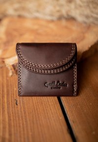 Portafoglio in pelle marrone scuro con dettagli di cucitura curvi e logo impresso "Gusti Leder studio", posato su una superficie di legno.