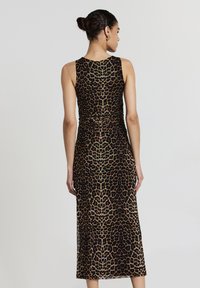 Femme avec les cheveux en chignon portant une robe sans manches, longue jusqu'aux chevilles, à motif léopard, tournée vers l'arrière contre un fond gris clair uni.