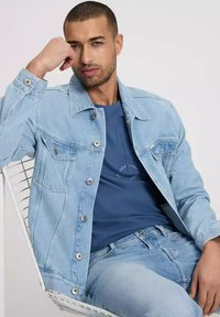 Jasnoniebieska kurtka dżinsowa z zapięciem na guziki i kieszeniami na piersi, nałożona na granatową koszulę, w zestawie z jasnoniebieskimi dżinsami.