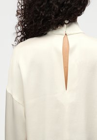 Blouse crème avec col montant et découpe en forme de goutte d'eau dans le dos. Texture douce et coupe ample avec des détails d'emmanchures tombantes.