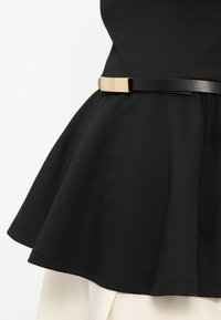 Μαύρο τοπ με καμπάνες, σφιγμένο στη μέση με λεπτή μαύρη ζώνη που έχει ορθογώνια χρυσή αγκράφα, σε συνδυασμό με λευκό παντελόνι.