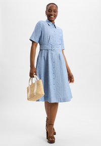 Blue chambray shirt dress with short sleeves, belted waist, button front, and flared skirt. Paired with a woven straw bag and platform heels.