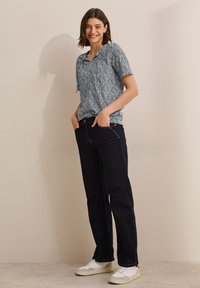 Blå mönstrad kortärmad blus kombinerad med mörka raka jeanstyg och vita sneakers. Blusen har en V-ringning och sidband.
