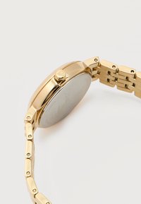 Ρολόι χειρός από μεταλλικό υλικό σε χρυσό τόνο με μπρασελέ από συνδεδεμένα κρίκους, απεικονιζόμενο από το πλάι, εστιάζοντας στις λεπτομέρειες της κορώνας και του κλειδώματος πάνω σε λευκό φόντο.