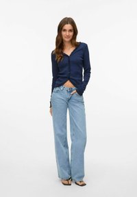 Marinblå långärmad cardigan med vågig nederkant, tillsammans med ljusblå vida jeans. Tyget är mjukt med en figursydd silhuett.