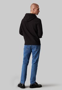 Sudadera negra con bolsillo canguro, combinada con jeans de mezclilla azules y zapatos negros. El diseño presenta un estilo simple y casual.