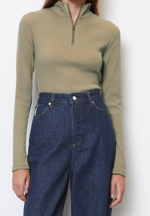 Femme portant un haut à manches longues côtelé vert olive avec fermeture éclair à l'avant et un jean en denim bleu foncé taille haute, sur fond blanc.