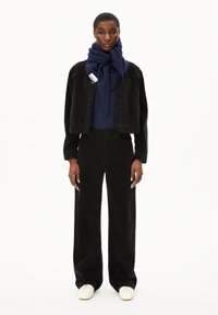 Schwarze Cordjacke, marineblauer Schal, dunkles Oberteil und weite schwarze Hose; beigefarbene Schuhe; glatte Textur, geschichteter Look, lässige Passform.