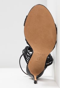 Chaussure à talon haut noire à brides, vue de la semelle avec un petit talon aiguille, appuyée contre un mur blanc.